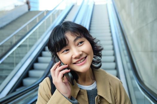 Close up portrait of smiling brunette girl, korean woman goes down escalator, makes phone call, talks to someone on telephone.