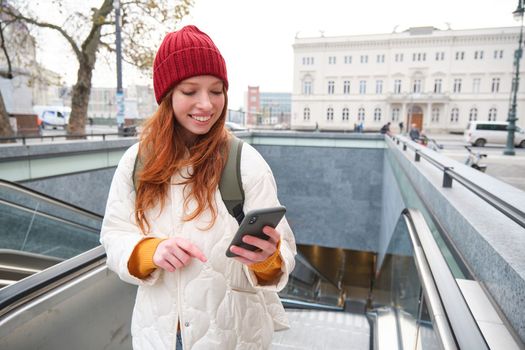 Portrait of young urban girl with red hair, using smartphone, texting message, using mobile phone application while going up an escalator.