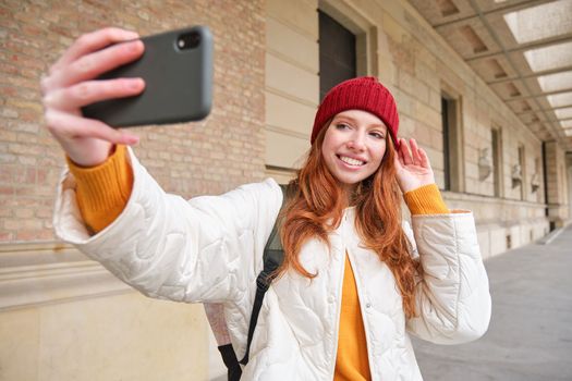 Stylish redhead girl tourist, takes selfie in front of tourism attraction, makes photo with smartphone, looks at mobile camera and poses.