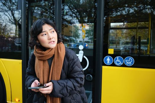 Korean girl in winter clothes, waits for her bus on stop, looking for her transport, holding mobile phone, checking timetable on smartphone application.