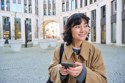 Stylish korean girl in headphones, listens music and uses mobile phone, stands in city centre, waits for someone on street and writes text message on her smartphone.