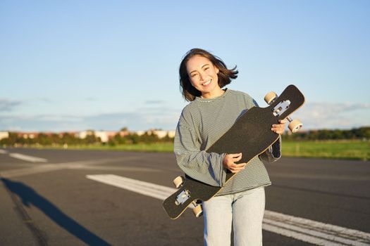 Lifestyle and hobbies. Smiling cute asian girl holding skateboard and walking towards sun on an empty road.