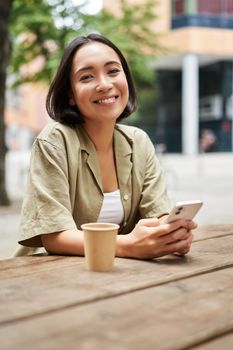 Portrait of young woman enjoying her coffee, drinking takeaway on bench in city, using smartphone. Copy space