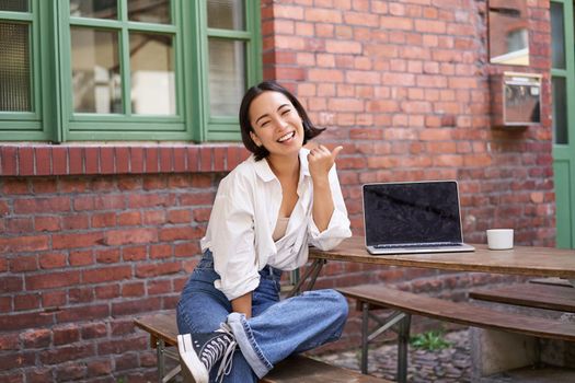 Happy korean girl with laptop, sitting in cafe, laughing and pointing at copy space on brick wall, showing advertisement.