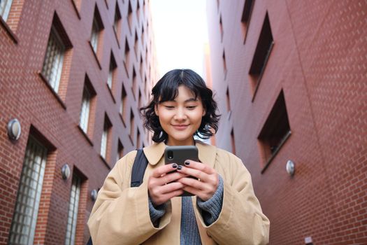 Lower angle view of brunette korean girl, listening music in headphones, walking along street and looking at smartphone, reading message on mobile phone.