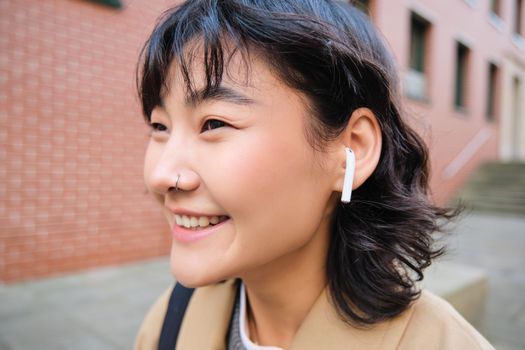 Walking around city. Smiling stylish korean girl in earphones, listens to music while walks on street, uses smartphone app, play songs.