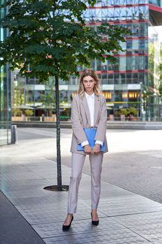 Portrait of young saleswoman in beige business suit, holding blue folder with work documents, standing outdoors on street of city center.