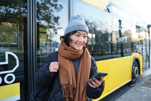 Portrait of girl standing near bus on a stop, waiting for her public transport, schecks schedule on smartphone application, holds mobile phone, wears warm clothes.