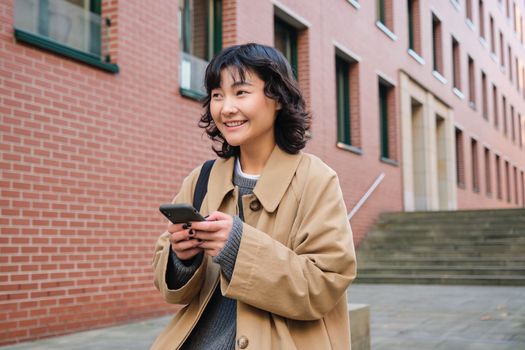 Stylish korean hipster girl, walks in city centre, sends text message, uses location map application on smartphone while walking down the street, wears trench coat.