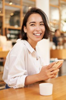 Portrait of young brunette woman, sitting with coffee and using smartphone in a cafe, chatting on mobile phone.