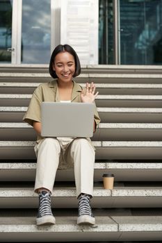 Portrait of young asian girl, student talks at laptop, video chat, speaking during online meeting, sitting outdoors on stairs.