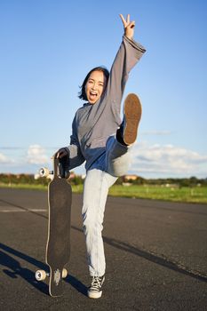 Excited asian girl dancing, standing with skateboard, skating on longboard and shouting carefree.