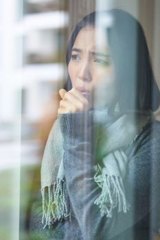 Portrait of sad ill korean woman looking out of the window, staying at home due to coronavirus, being sick and cant leave house.