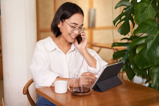 Woman in glasses, answer phone call in a cafe, drinking coffee, looking at work on tablet, working on remote from restaurant.