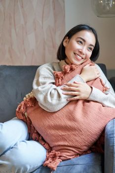 Portrait of asian woman feeling cozy in her home, hugging pillow on sofa and looking thoughtful, smiling.