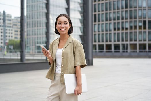 Asian girl with laptop and smartphone, standing on street of city centre, smiling at camera.