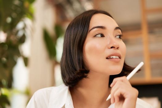 Close up portrait of stylish young brunette woman, sitting with graphic pen and smiling, relaxing in cafe, writing something, making notes.