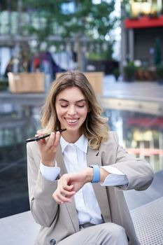 Vertical shot of smiling woman records voice message and looks at digital watch, reads message or checks time, sits in city centre.