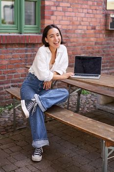 Vertical shot of good-looking asian woman with laptop, sitting in outdoor cafe, drinking coffee and smiling, laughing carefree.