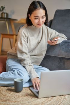 Vertical shot of young woman in cozy home working on laptop, using smartphone and drinking coffee, sitting on floor near sofa.