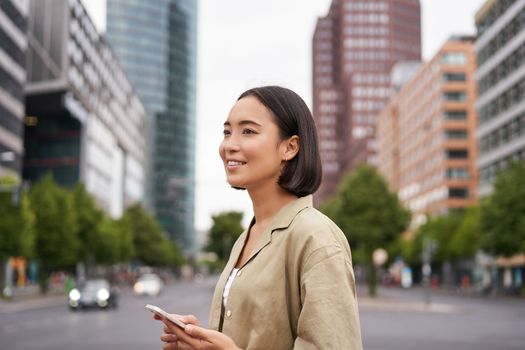 Young asian woman exploring city with smartphone app, holding mobile phone and walking on screet.