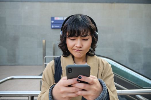 Portrait of asian girl in headphones, looks complicated at smartphone screen, puzzled by text message or notification, stands on street and shrugs.