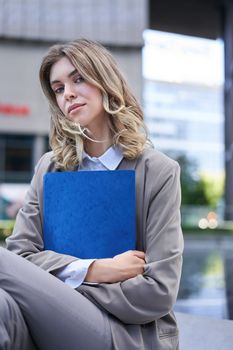 Corporate woman holding documents in hands, sitting on a street, wearing office suit, looking confident at camera, carry a folder with work.