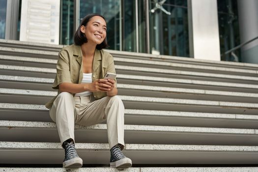 Portrait of happy smiling asian woman, sitting outdoors near building, using smartphone. Technology concept.