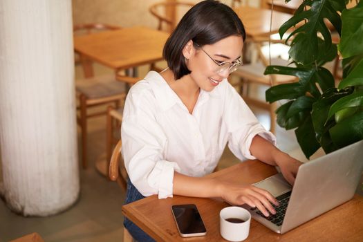 Working female entrepreneur sitting in cafe with laptop, typing on computer, drinking coffee.