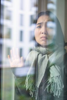 Asian woman feels unwell, stays at home, looks outside window with jealous, distressed face, wants go outside but caught cold.