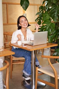 Happy young asian woman winning on laptop, receive good news, achieve goal at work, triumphing and smiling pleased, sitting in cafe.