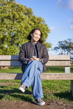 Vertical shot of beautiful brunette woman sitting on bench with smartphone, waiting for someone, using mobile appliation.