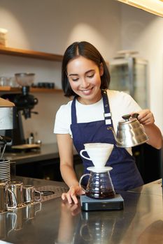 Vertical shot of cute asian girl barista, student working in coffee shop, pouring water in filter. brewing, standing in apron behind counter in cafe.