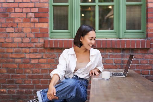 Portrait of young stylish woman, influencer sitting in cafe with cup of coffee and laptop, smiling and looking confident.