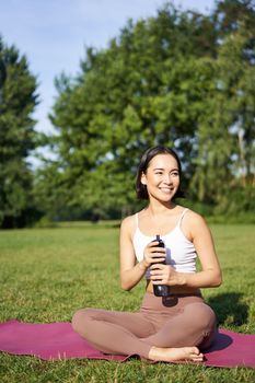 Smiling fitness girl sits on mat and drinks water after workout, finish yoga training in park, exercise on fresh air.