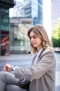 Frowning businesswoman in suit, looking at her digital watch with disappointed face, annoyed waiting for someone, sitting outside.