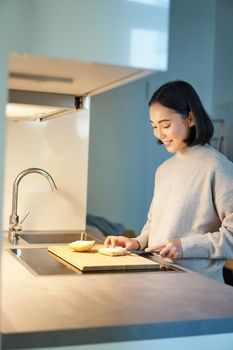 Vertical shot of young asian woman cooking dinner, making herself sandwitch, smiling while standing on the kitchen.