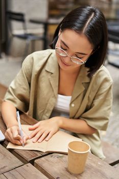 Vertical shot of young asian woman doing homework, making notes, writing something down, sitting in an outdoors cafe and drinking coffee.