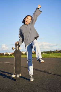 Excited asian girl dancing, standing with skateboard, skating on longboard and shouting carefree.