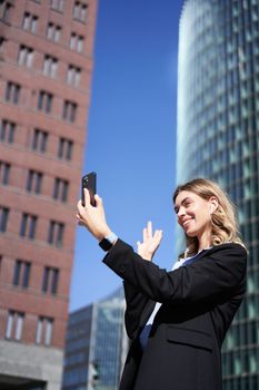 Portrait of smiling corporate woman video call on street, holding mobile phone and waving at smartphone camera, wearing suit.