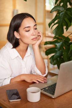 Vertical shot of asian girl looking tired at laptop screen, bored of working, sitting in cafe sleepy and exhausted.