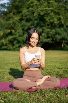 Portrait of korean girl meditating in park, follow yoga video lesson on smartphone app, sitting on fresh air and relaxing.