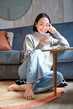 Vertical shot of stylish korean woman sitting on floor at home, using her mobile phone, holding smartphone and smiling.