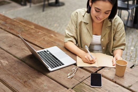 Portrait of young woman studying online, sitting with laptop, writing down, making notes and looking at computer screen, sitting in cafe outdoors.