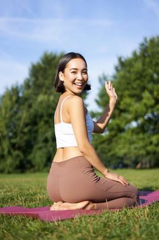 Portrait of asian girl on fitness class in park, sitting on rubber mat and wave hand at camera, say hello, meditating and practice yoga.