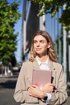 Vertical shot of successful business woman, team leader in suit, holding digital tablet, walking on street. Corporate lady goes to work.