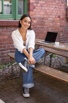 Stylish young businesswoman, asian girl with laptop, sitting in outdoor cafe with cup of coffee and working, using computer.