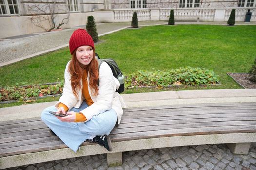 Young stylish female student with backpack, sits on bench and uses smartphone, plays on mobile phone.