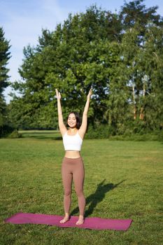 Vertical shot of asian woman raising hands up to sky, meditating, practice yoga, doing wellbeing training in park on green lawn.