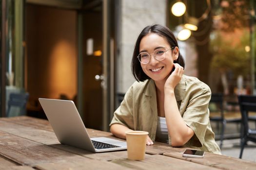 Portrait of smiling girl in glasses, sitting with laptop in outdoor cafe, drinking coffee and working remotely, studying online.